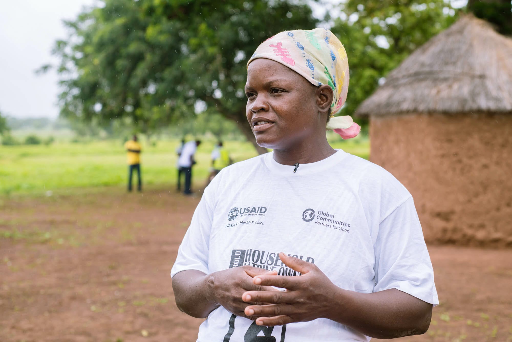 Woman wearing a GC-USAID branded shirt with the inscription, "I AM A NATURAL LEADER", with a mud house behind her